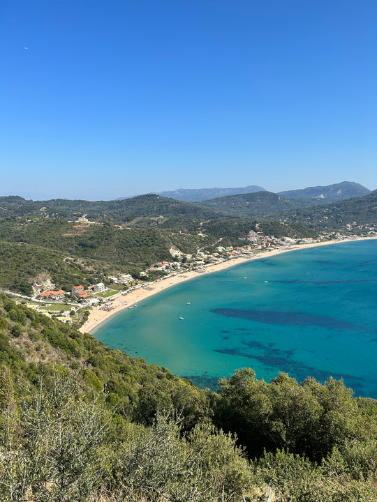 Stunning view from a mountain on Corfu Island, showcasing the crystal-clear turquoise waters of the Ionian Sea and a picturesque beach nestled along the coastline, surrounded by lush greenery.